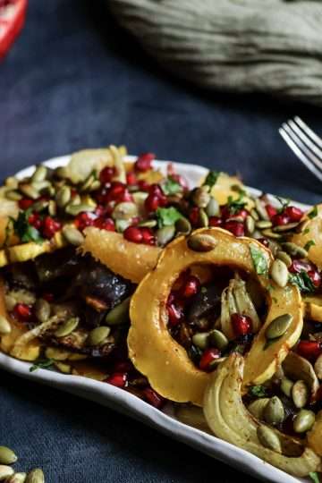 White rectangular platter filled with roasted delicata squash and fennel salad topped with pomegranate seeds, set on a dark grey background. A halved pomegranate and a small white bowl of pomegranate seeds sit nearby, with a fork, an olive-green cloth, and a few scattered pumpkin seeds in the background.