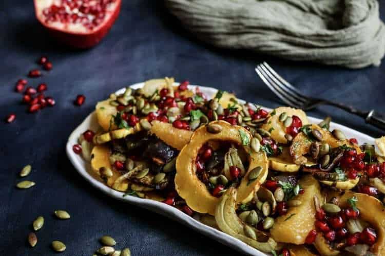 White rectangular platter filled with roasted delicata squash and fennel salad topped with pomegranate seeds, set on a dark grey background. A halved pomegranate and a small white bowl of pomegranate seeds sit nearby, with a fork, an olive-green cloth, and a few scattered pumpkin seeds in the background.