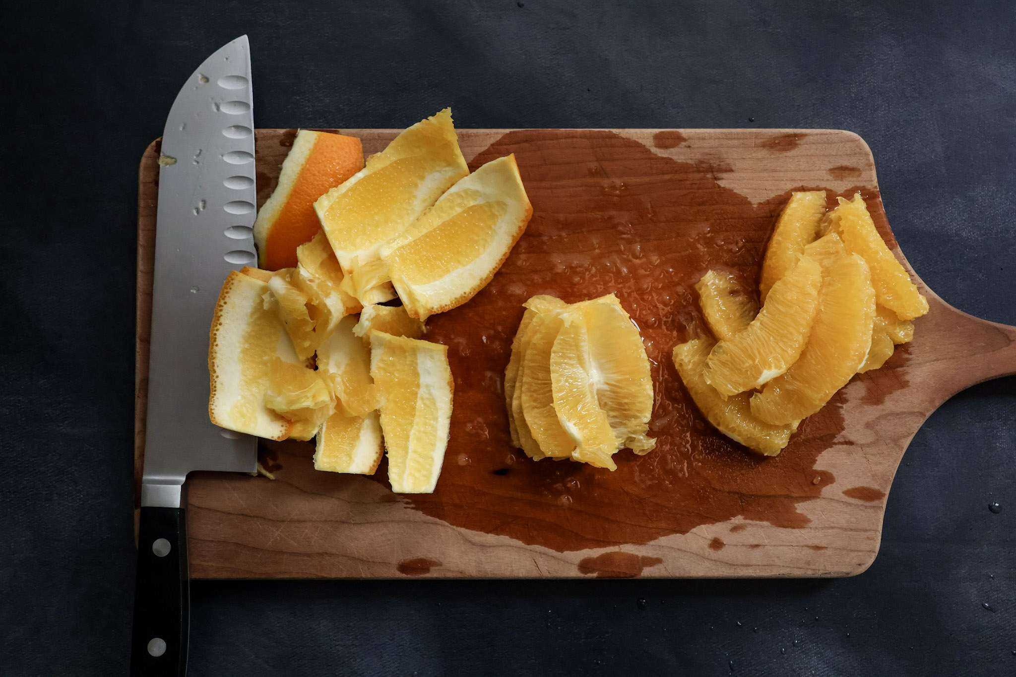 Wooden board on a dark grey surface holding a chef’s knife, strips of orange peel, segmented orange slices, and the remaining orange core after the segments were removed.