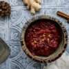 A small bowl of cranberry chia jam with orange and ginger is in the center, surrounded by a couple pine cones, two cinnamon sticks, a frayed beige napkin and a small silver candleholder
