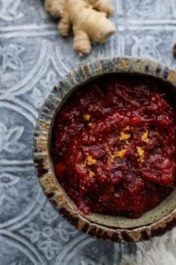 A small bowl of cranberry chia jam with orange and ginger is in the center, surrounded by a couple pine cones, two cinnamon sticks, a frayed beige napkin and a small silver candleholder