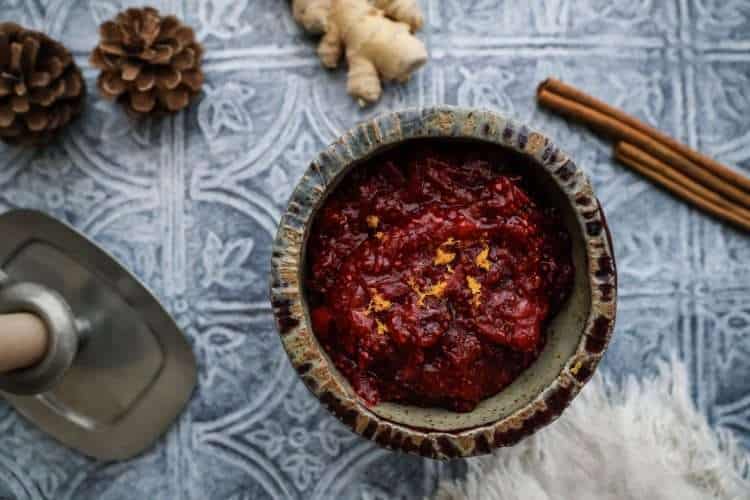 A small bowl of cranberry chia jam with orange and ginger is in the center, surrounded by a couple pine cones, two cinnamon sticks, a frayed beige napkin and a small silver candleholder