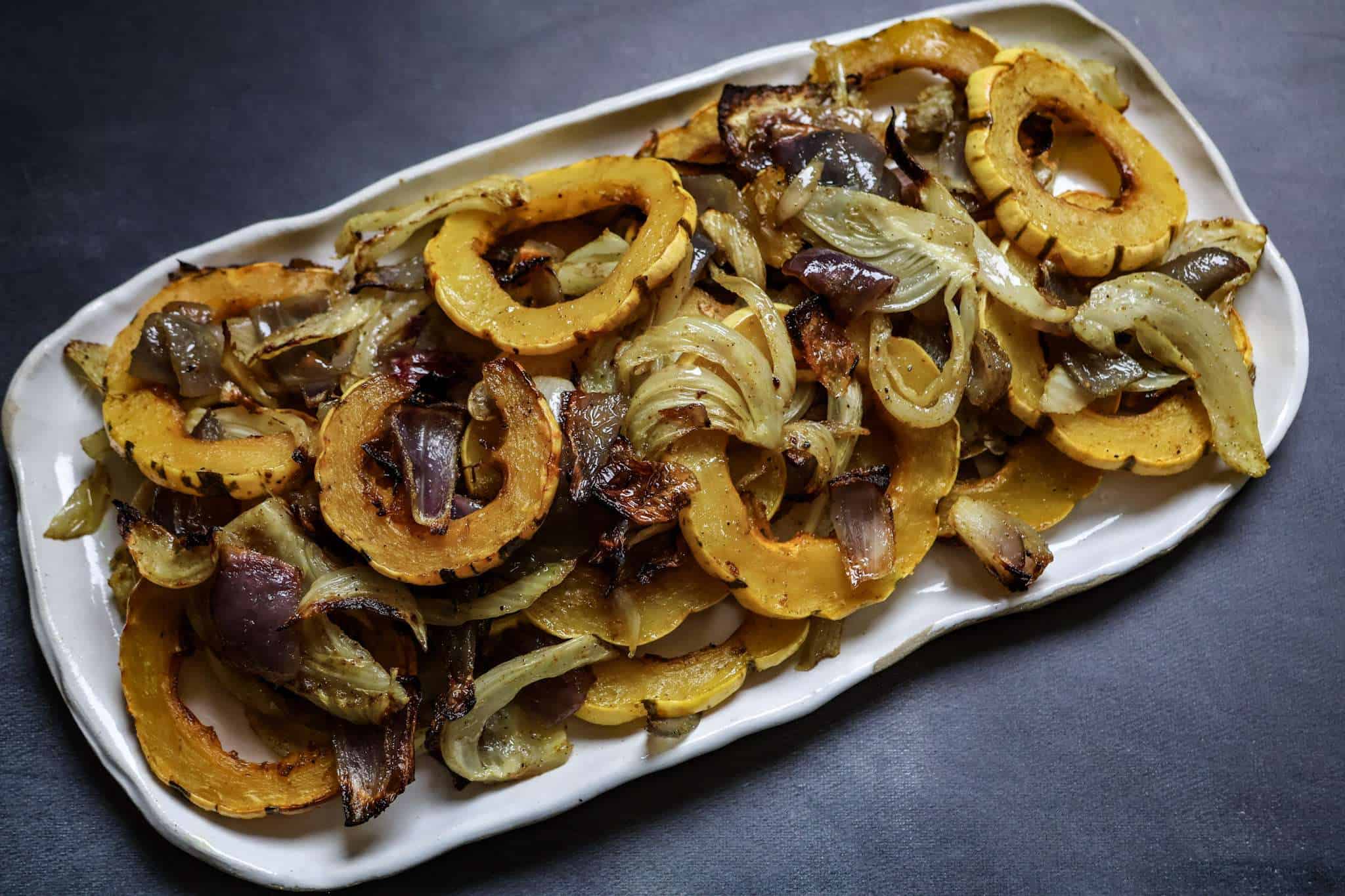 White rectangular platter filled with Ras el Hanout–seasoned roasted delicata squash, fennel, and red onions on a dark gray background.