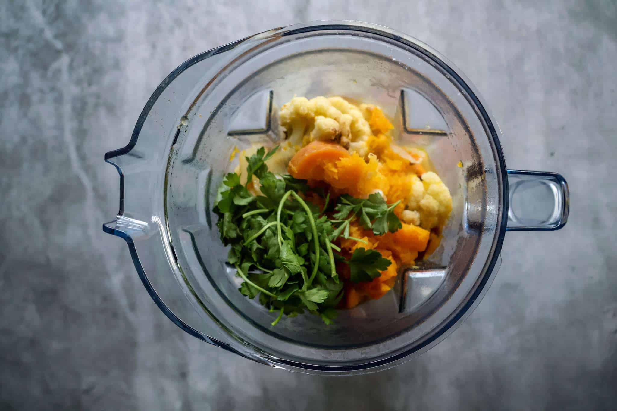 a blender container with roasted vegetables, broth and parsley for creamy winter vegetables soup