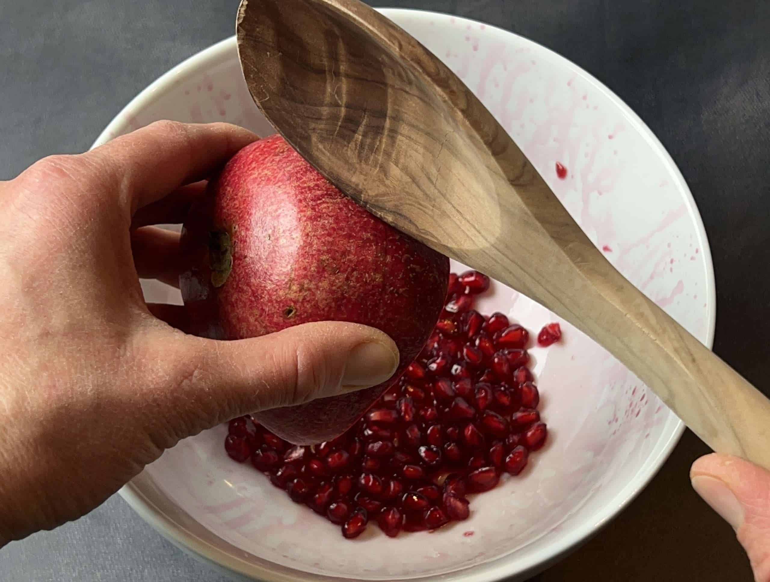White bowl filled with pomegranate seeds, a hand holding half a pomegranate, and another hand tapping the back of the pomegranate with a wooden spoon to release the seeds.