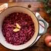 A pot of nourishing spiced red cabbage with two thin apple slices and a cinnamon stick on top, a cream colored napkin with red stitching is wrapped around the pot, an apple it placed in the lower right hand corner, a star anise is is placed on the wooden surface above the pot and some evergreen sprigs can be seen on the right hand side