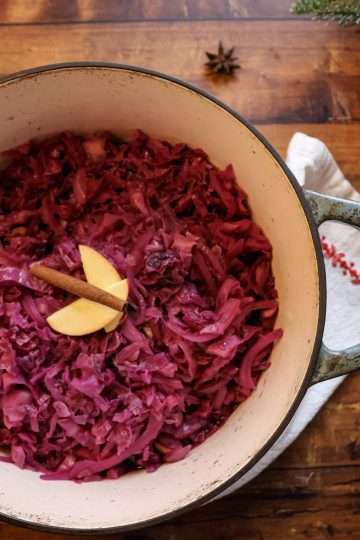A pot of nourishing spiced red cabbage with two thin apple slices and a cinnamon stick on top, a cream colored napkin with red stitching is wrapped around the pot, an apple it placed in the lower right hand corner, a star anise is is placed on the wooden surface above the pot and some evergreen sprigs can be seen on the right hand side
