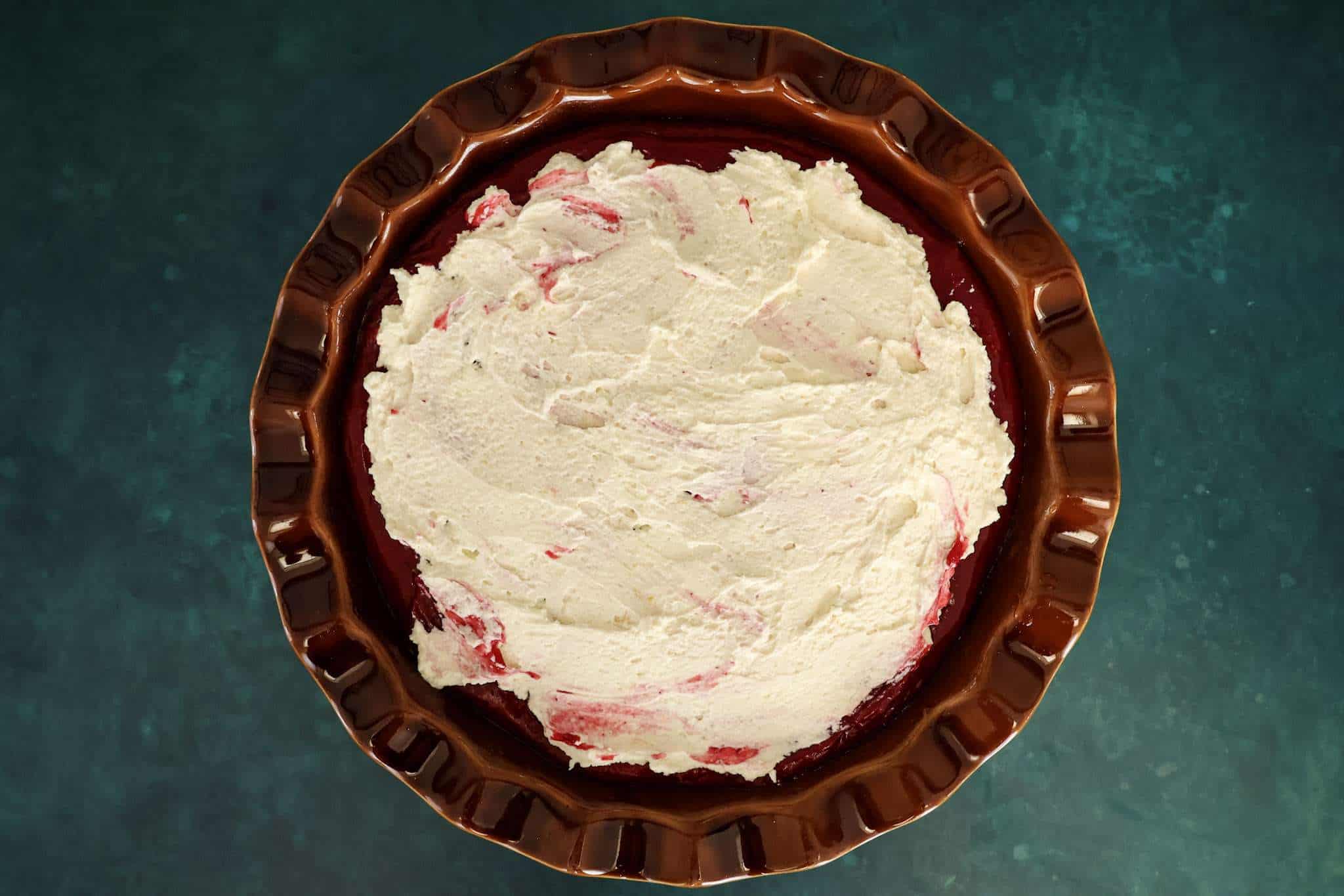 A brown pie dish with the cranberry orange tiramisu pie in progress, showing a thin, uneven layer of mascarpone frosting spread over the cranberry purée like a crumb coat