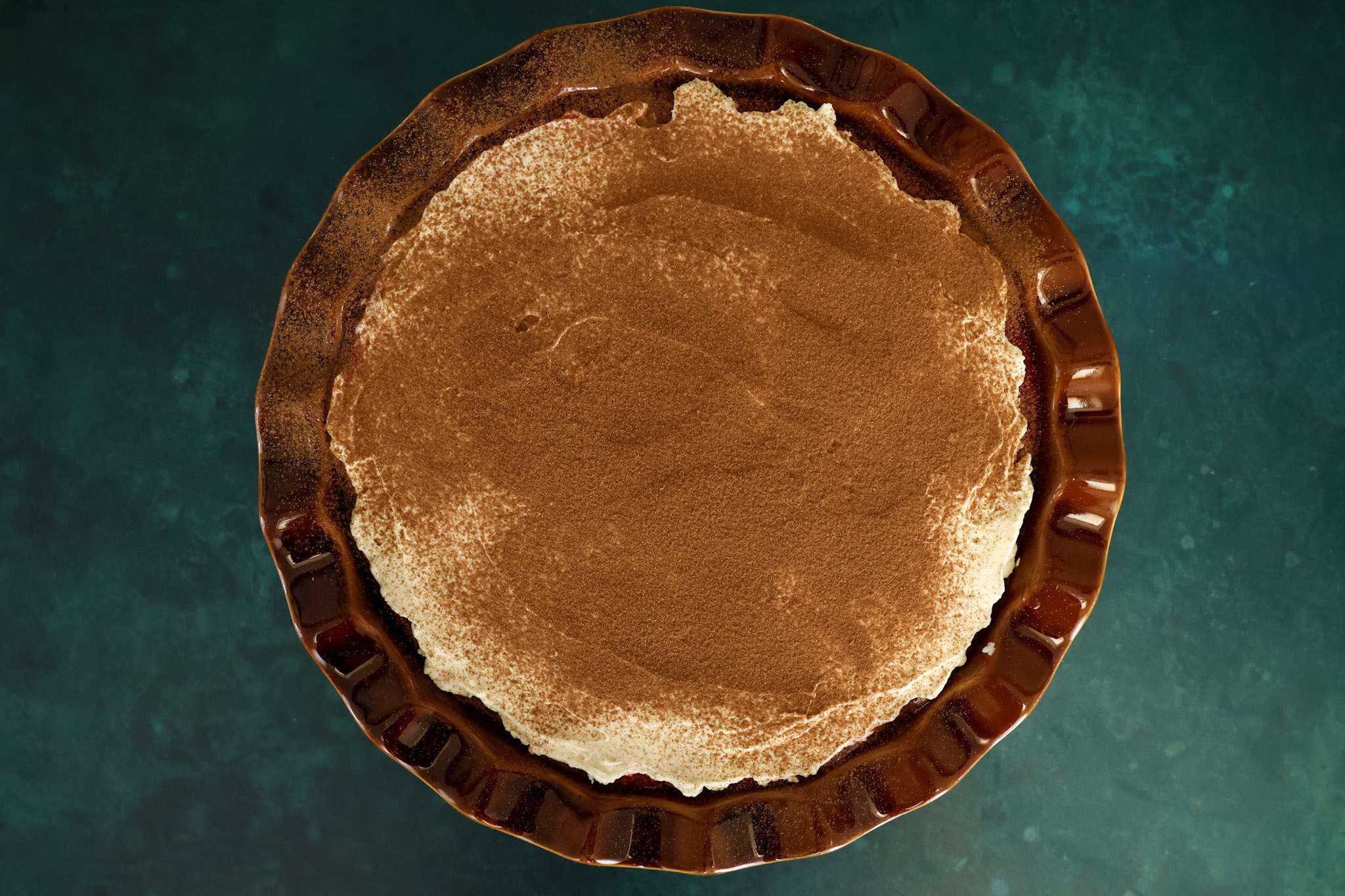 A brown pie dish filled with the finished cranberry orange tiramisu pie, topped with a smooth, even layer of mascarpone-style frosting and a light dusting of cinnamon