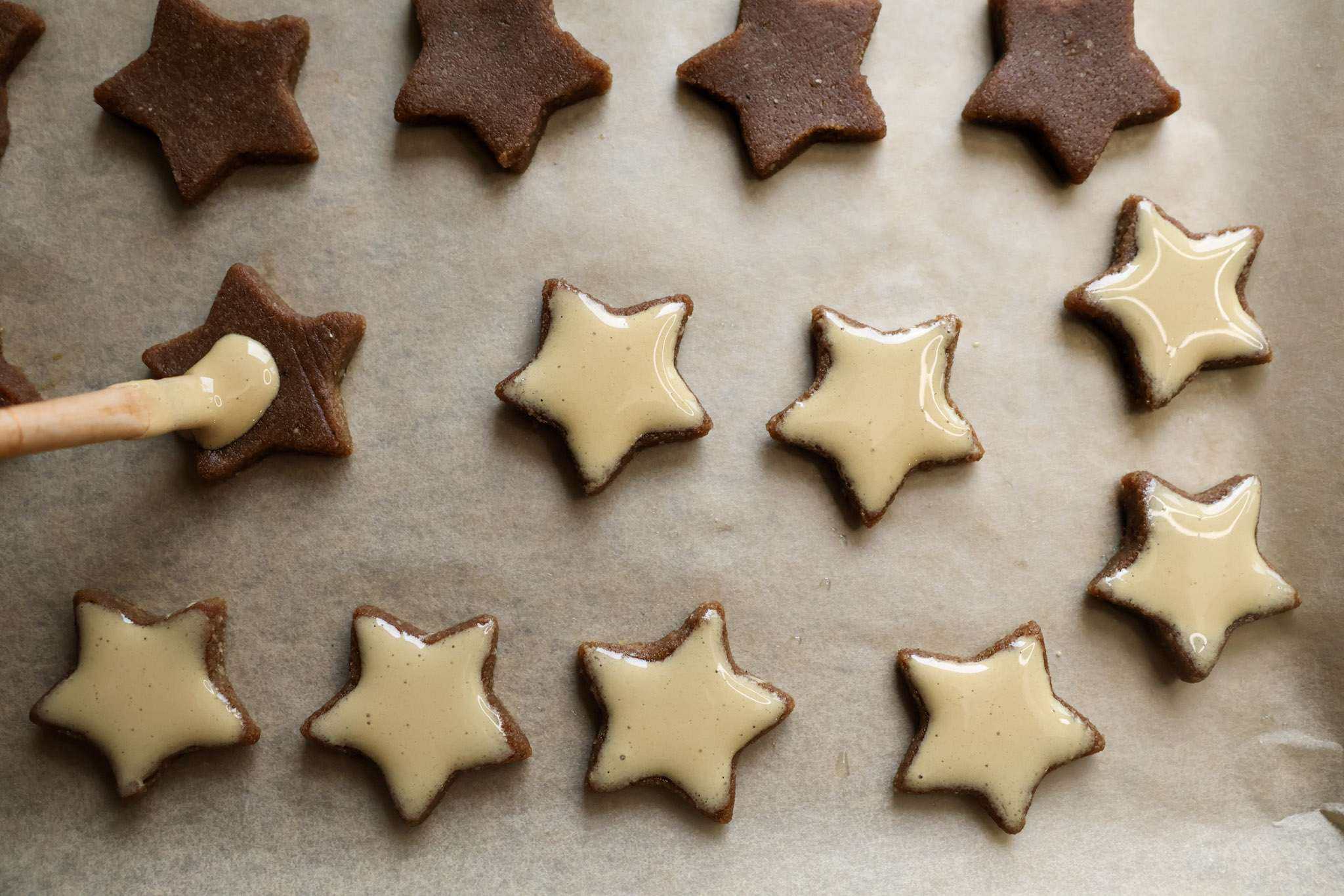 A cookie sheet with zimsterne cookies on it - some are already glazed with their meringue top, some are still waiting to be glazed and one is being glazed with a brush