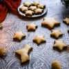 German Zimtsterne (cinnamon star) cookies are scattered on a textured grey surface - a German style wooden candle holder is pictured in the background along with an additional small plate of cookies, a few whole almonds in the front and a red cloth and some evergreen branches can be seen in the background