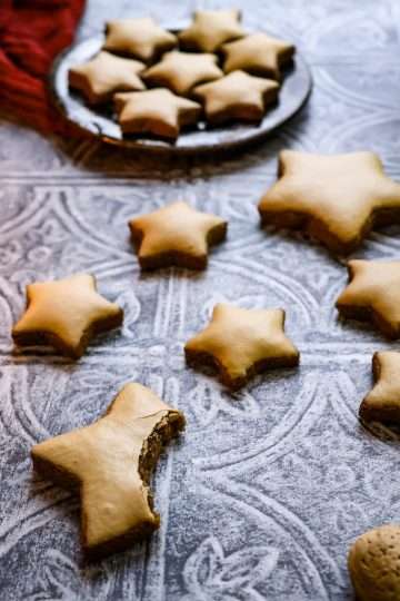 German Zimtsterne (cinnamon star) cookies are scattered on a textured grey surface - a German style wooden candle holder is pictured in the background along with an additional small plate of cookies, a few whole almonds in the front and a red cloth and some evergreen branches can be seen in the background