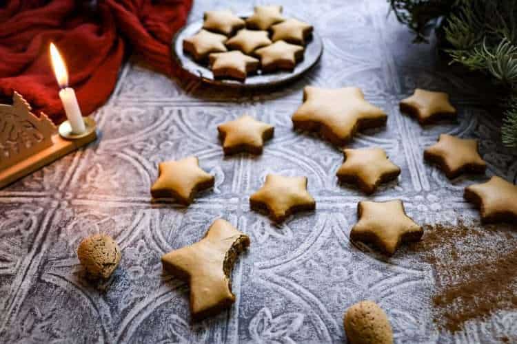German Zimtsterne (cinnamon star) cookies are scattered on a textured grey surface - a German style wooden candle holder is pictured in the background along with an additional small plate of cookies, a few whole almonds in the front and a red cloth and some evergreen branches can be seen in the background