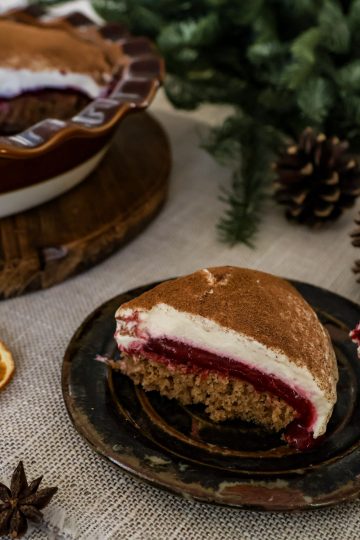 A slice of cranberry orange tiramisu pie on a small rustic plate with a spoonful beside it, with a star anise pod and dehydrated orange slice on the tablecloth in front, pine cones and greenery in the background, and the remaining pie in a dish on a wooden board behind it.