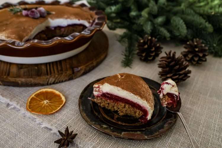 A slice of cranberry orange tiramisu pie on a small rustic plate with a spoonful beside it, with a star anise pod and dehydrated orange slice on the tablecloth in front, pine cones and greenery in the background, and the remaining pie in a dish on a wooden board behind it.