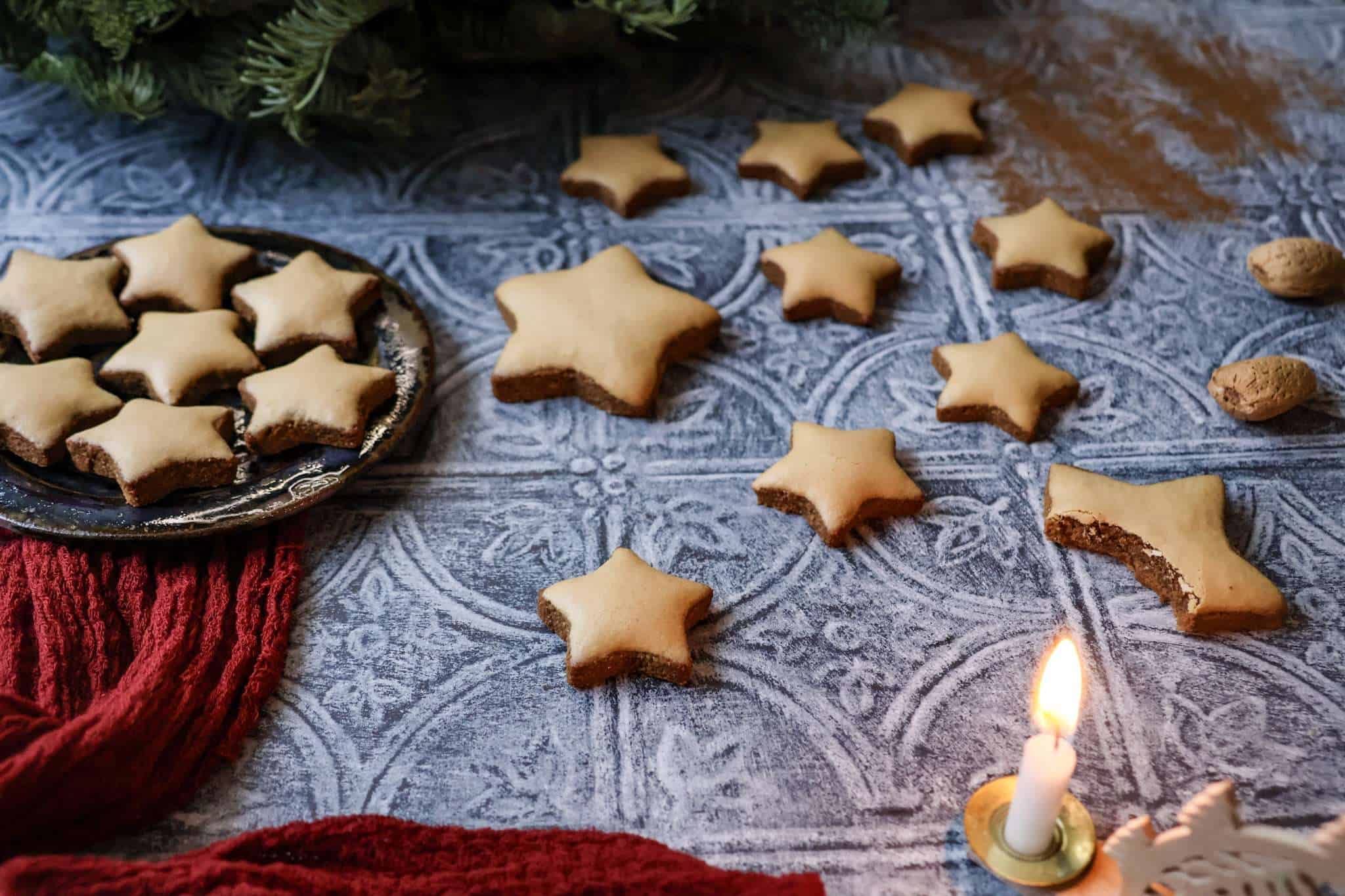 German Zimtsterne (cinnamon star) cookies are scattered on a textured grey surface - a German style wooden candle holder is partially pictured in the forefront and an additional small plate of cookies can be seen to the left and a red cloth and some evergreen branches can be seen as well