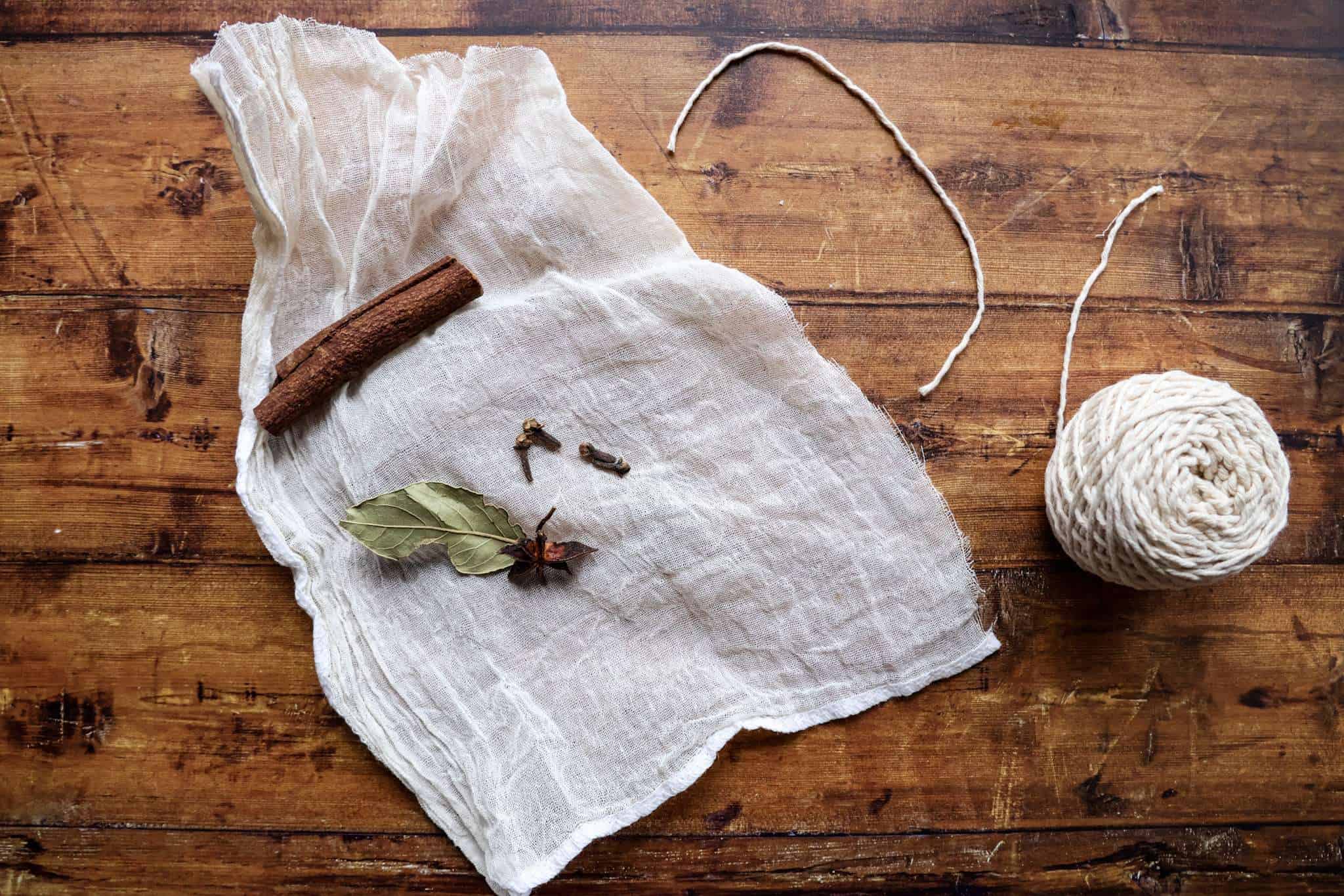 a cheese cloth with a cinnamon stick, cloves, bay leaf, and star anise is placed on a wooden surface with kitchen twine next to it