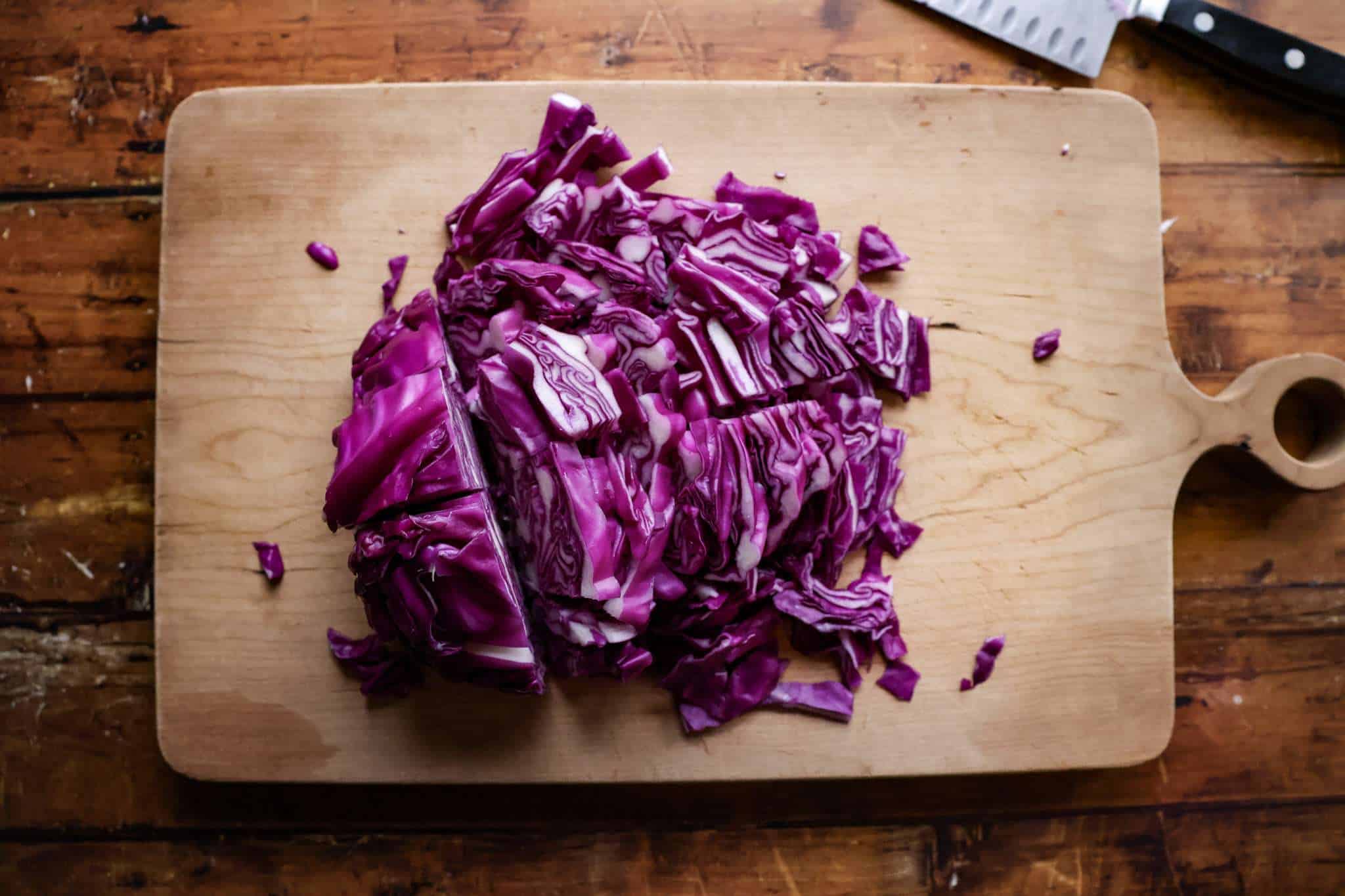 half a head of red cabbage partially sliced thinly on a wooden board with a chef knife resting behing the board