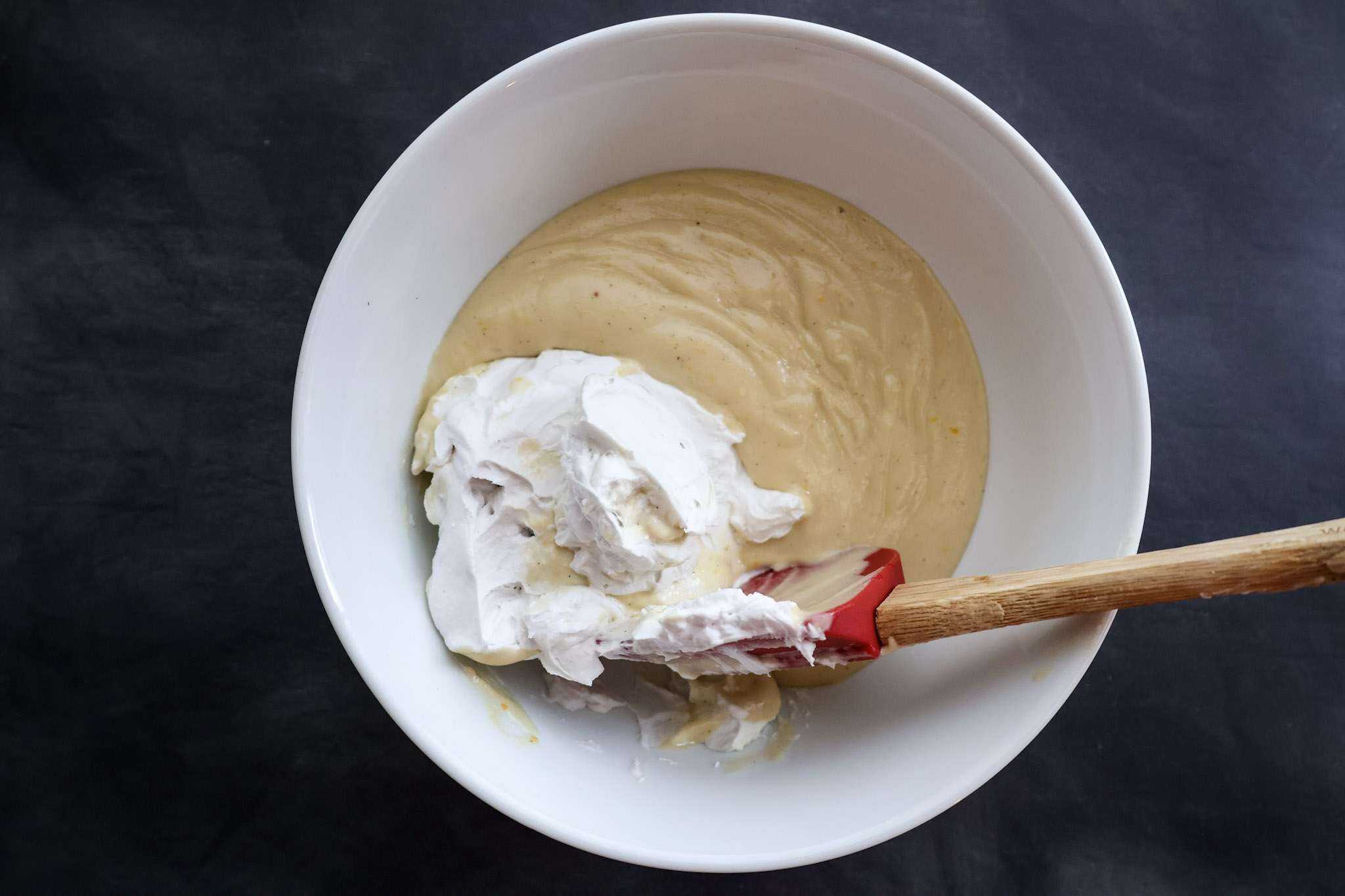 Cashew cream for dairy-free mascarpone style frosting in a while bowl and whipped coconut cream being fold in with a red spatula