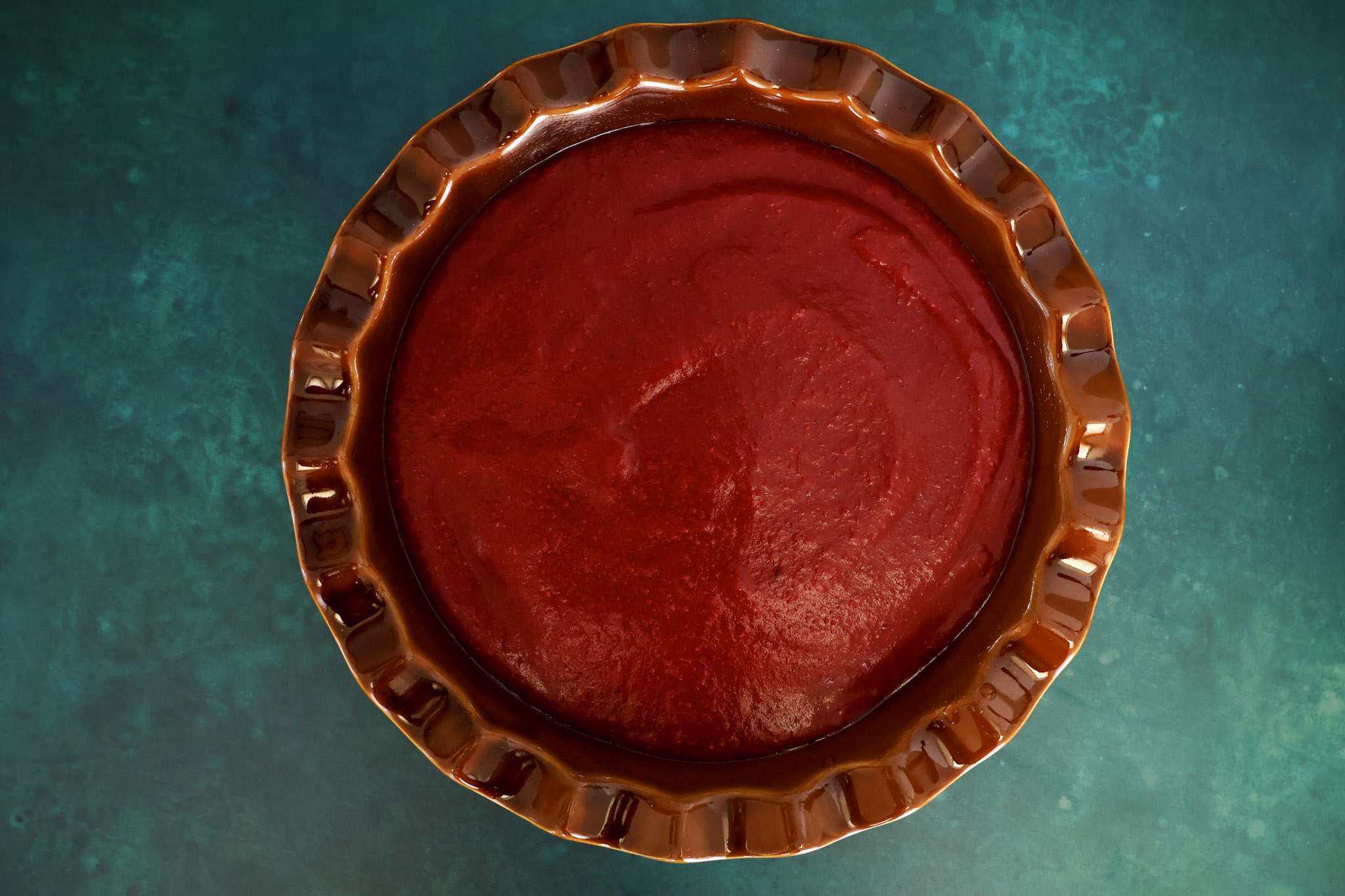 A brown pie dish holding the partially assembled cranberry orange tiramisu pie, showing only the smooth cranberry purée layer spread over the sponge before the cream layer is added