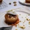 In the forefront is a white plate with a small orange walnut frangipane style cake sitting in a small pool of quick orange jam and topped with a quenelle of coconut creme fraiche and a sprinkle of finely chopped walnuts - a black fork is resting on the plate and another similar plate is in the background, along with a cooling rack holding additional small cakes.