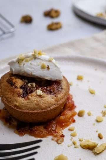 In the forefront is a white plate with a small orange walnut frangipane style cake sitting in a small pool of quick orange jam and topped with a quenelle of coconut creme fraiche and a sprinkle of finely chopped walnuts - a black fork is resting on the plate and another similar plate is in the background, along with a cooling rack holding additional small cakes.
