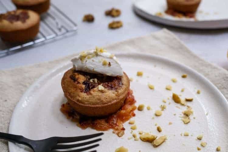 In the forefront is a white plate with a small orange walnut frangipane style cake sitting in a small pool of quick orange jam and topped with a quenelle of coconut creme fraiche and a sprinkle of finely chopped walnuts - a black fork is resting on the plate and another similar plate is in the background, along with a cooling rack holding additional small cakes.