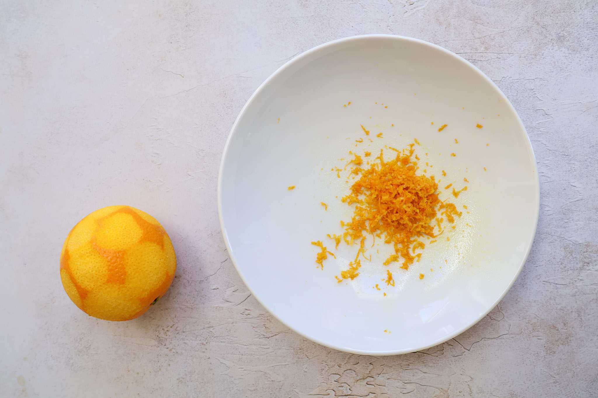 A shallow white bowl holding orange zest and a zested orange sitting off to the side