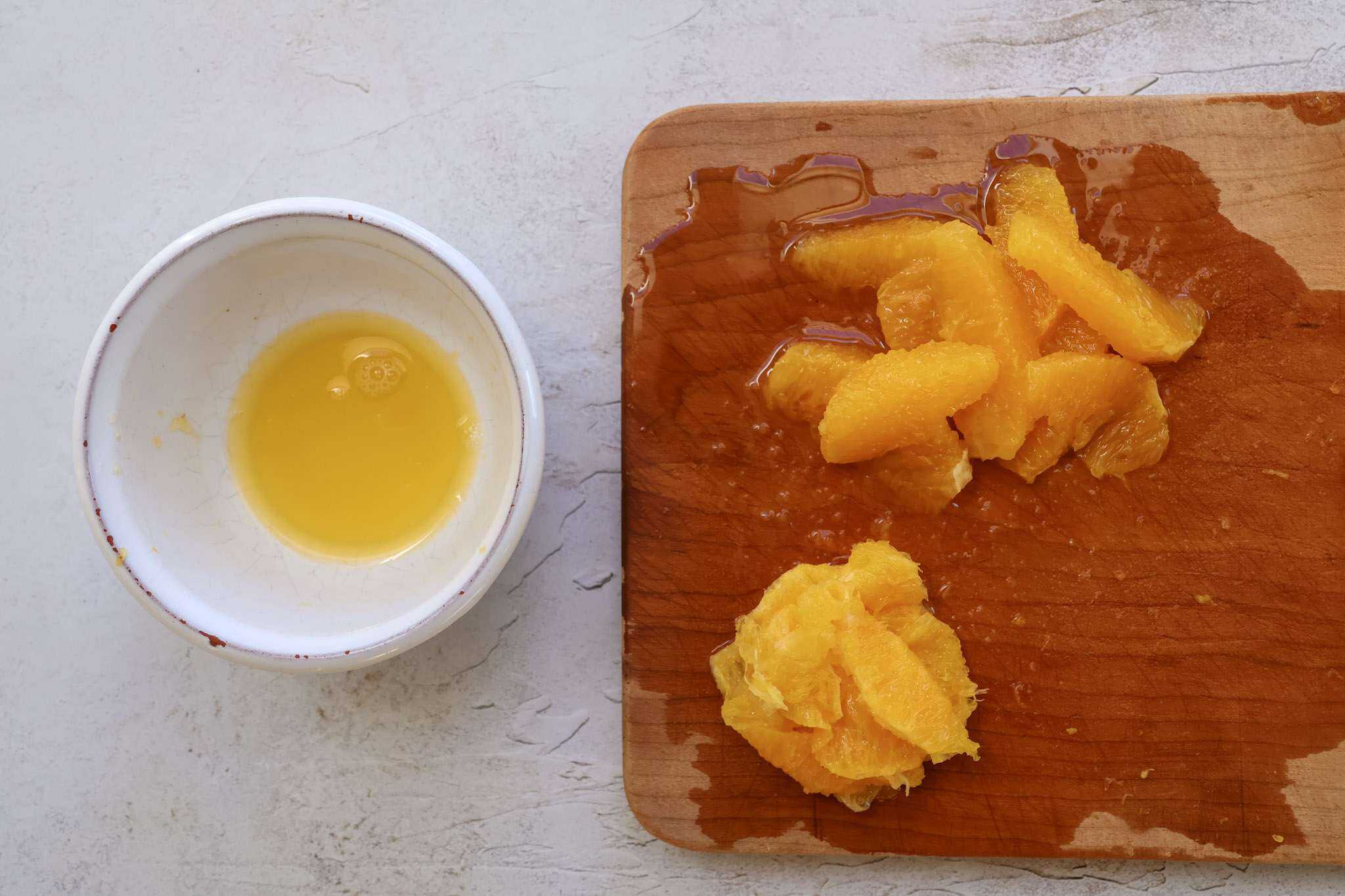 A small white bowl with orange juice is on the left side and on the right side is a wooden cutting board with segmented orange slices and the core of a segmented orange