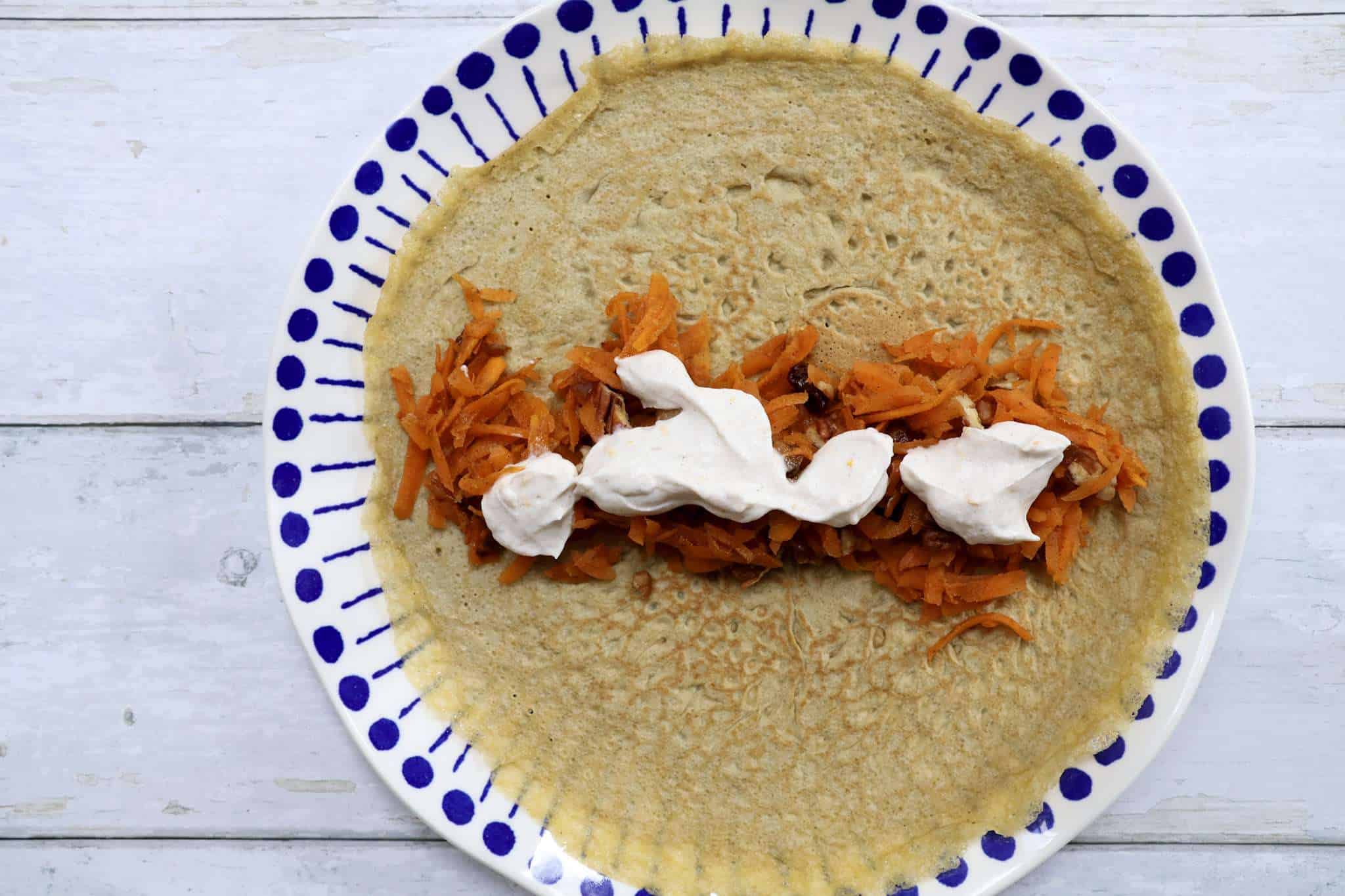 A crepe topped with carrot cake crepe filling and coconut yogurt topping on a white and blue plate