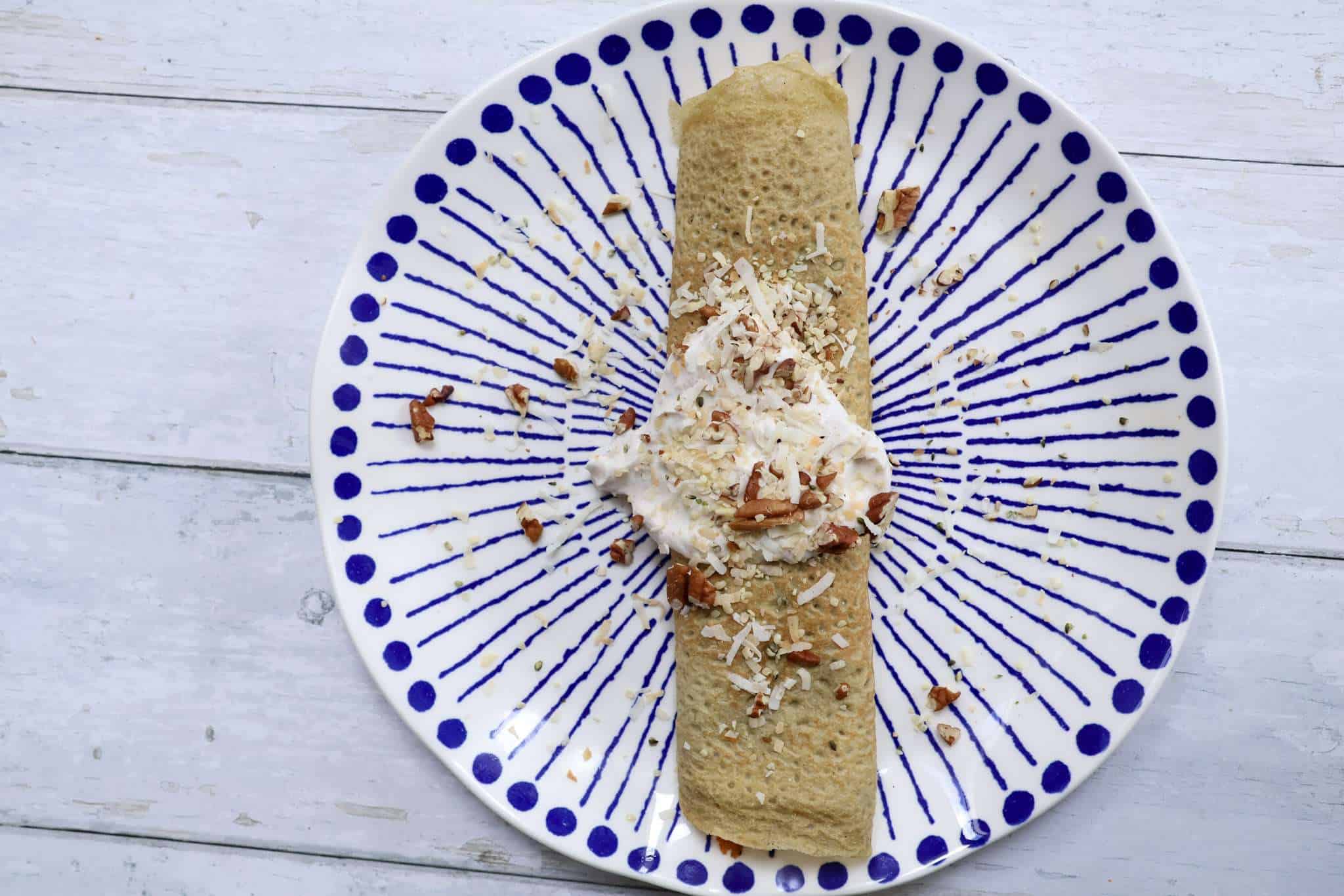 A fully assembled carrot cake crepe, topped with coconut yogurt, chopped toasted pecans, toasted shredded coconut, and hemp seeds on a white and blue plate