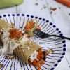 A blue and white plate topped with a carrot cake crepe - a black fork is resting on the plate, a green napkin is in the background and some carrots and chopped pecans are scattered in the background along with a small bowl with coconut yogurt