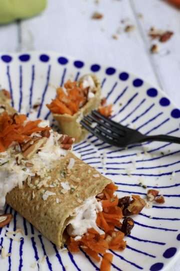 A blue and white plate topped with a carrot cake crepe - a black fork is resting on the plate, a green napkin is in the background and some carrots and chopped pecans are scattered in the background along with a small bowl with coconut yogurt