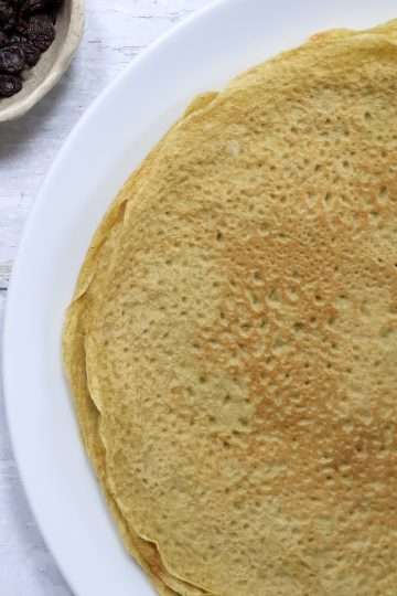 A white plate with crepes and small bowls of maple syrup and raisins are off to the left side