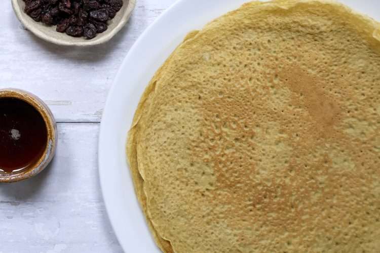A white plate with crepes and small bowls of maple syrup and raisins are off to the left side