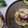 A rustic dark colored plate on a concrete style surface - the plate is topped with a grain-free tortilla with sheet pan chicken and onions, spiced rhubarb chutney, a dollop of coconut yogurt, a sprinkle of cilantro, and radish sliced matchstick size. A tortilla warmer can be seen in the bottom right side of the picture and a small bundle of cilantro is also in the background