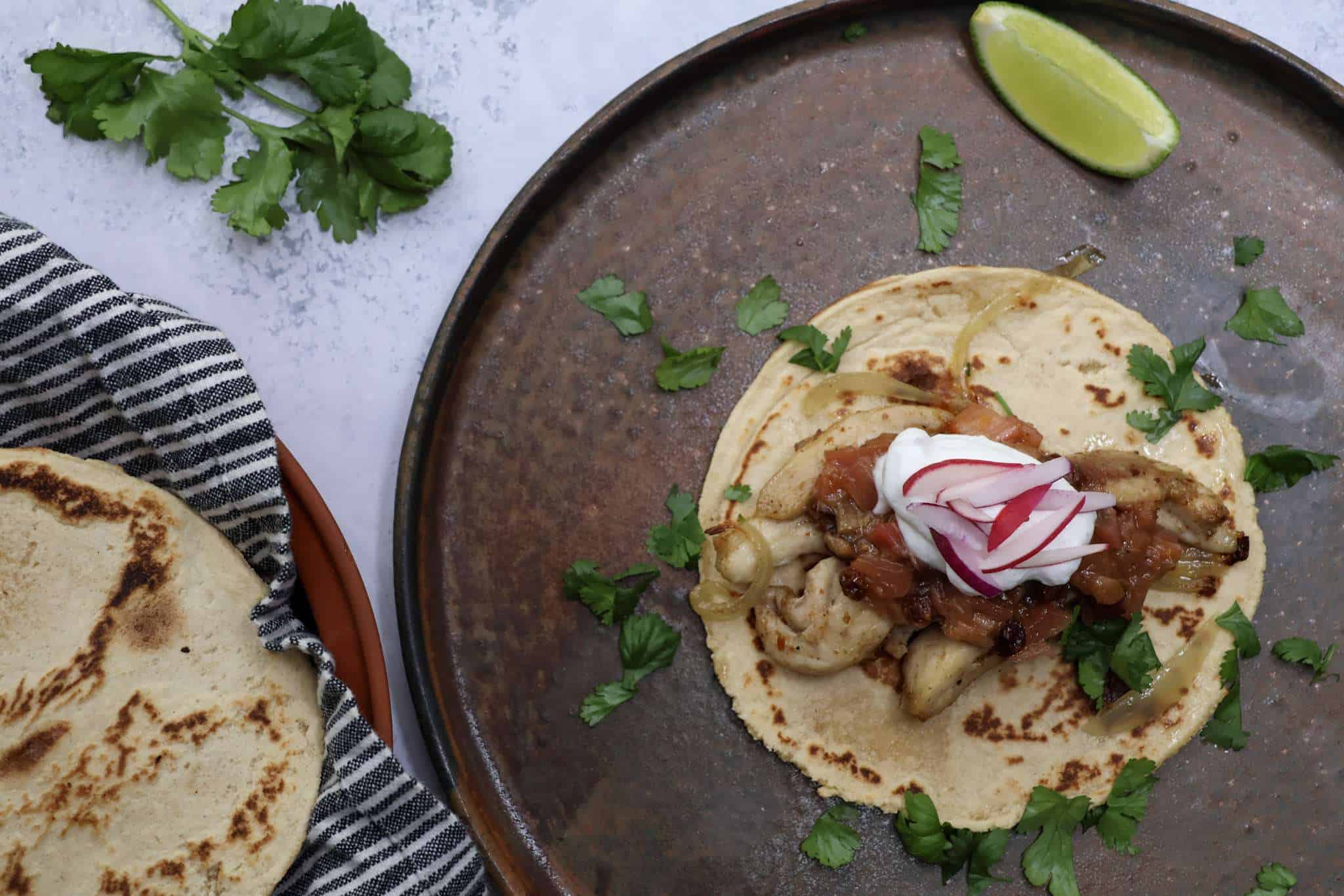 A rustic dark colored plate on a concrete style surface - the plate is topped with a grain-free tortilla with sheet pan chicken and onions, spiced rhubarb chutney, a dollop of coconut yogurt, a sprinkle of cilantro, and radish sliced matchstick size. A tortilla warmer can be seen in the bottom right side of the picture and a small bundle of cilantro is also in the background