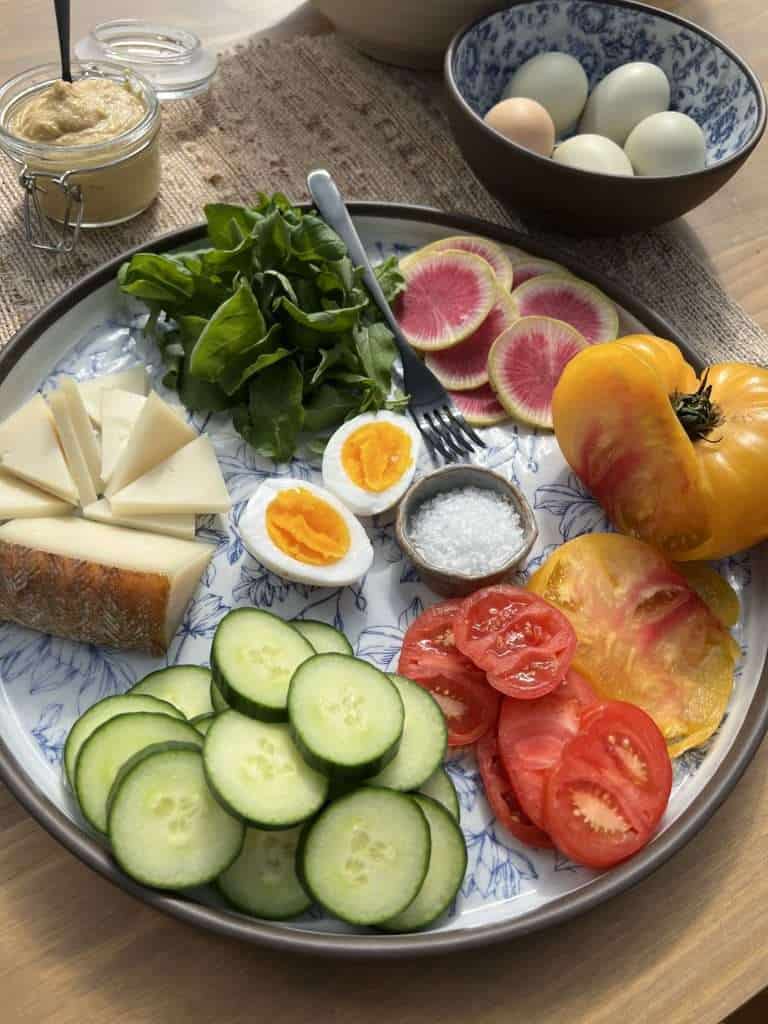 a beautiful blue and white flower platter with sliced veggies, cheese and a boiled egg and a chickpea dip and boiled eggs in the background