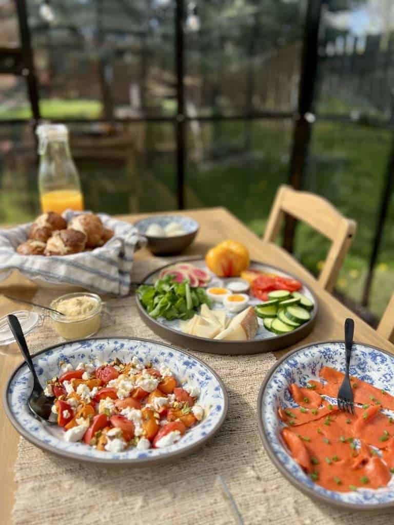 a breakfast table set in a greenhouse with a bottle of orange juice, blue and white platters with grilled apricots, goat cheese, sliced vegetables, smoked salmon, boiled eggs and a bowl with pretzel rolls