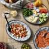 Summer brunch spread on a rustic wooden table with blue platters of smoked salmon, vegetables, cheese, olives, chive blossom butter, and grilled apricots with goat cheese, pistachios, and honey.