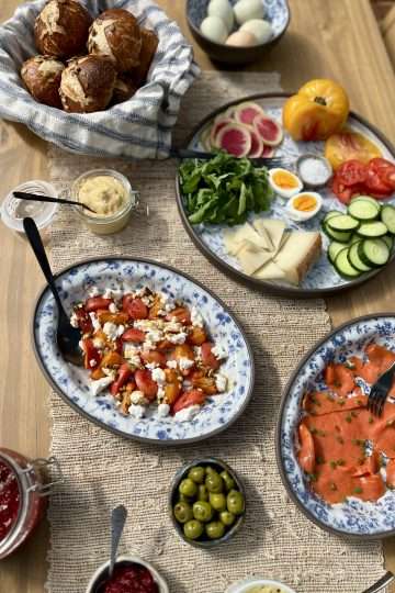 Summer brunch spread on a rustic wooden table with blue platters of smoked salmon, vegetables, cheese, olives, chive blossom butter, and grilled apricots with goat cheese, pistachios, and honey.