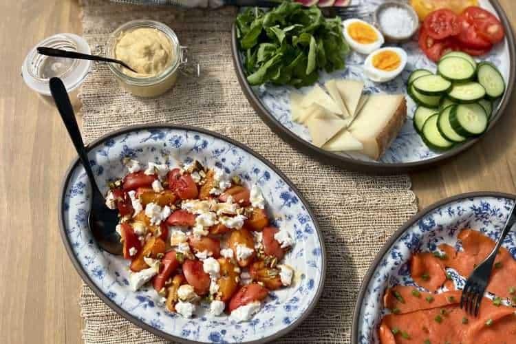 Summer brunch spread on a rustic wooden table with blue platters of smoked salmon, vegetables, cheese, olives, chive blossom butter, and grilled apricots with goat cheese, pistachios, and honey.