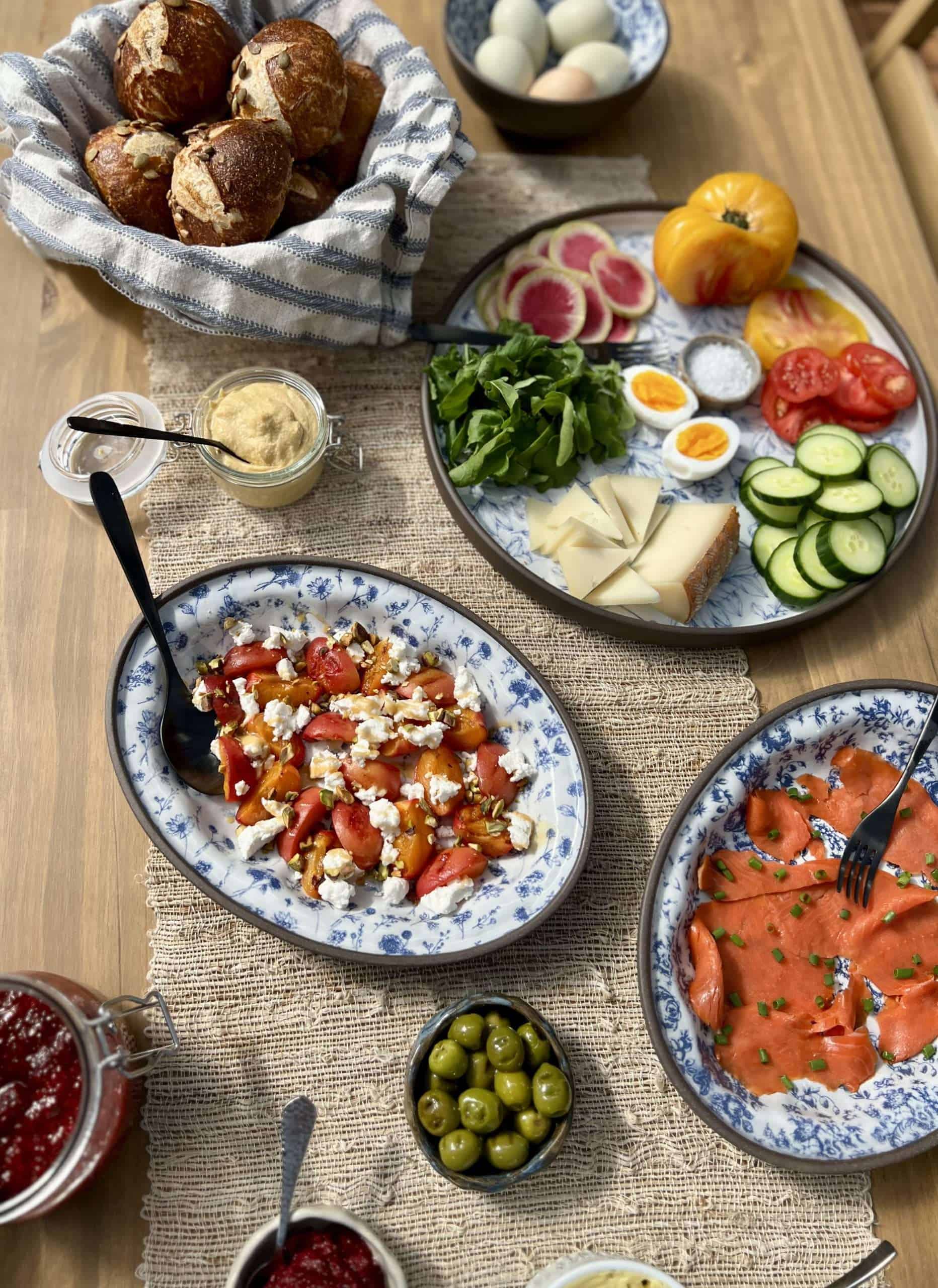 Summer brunch spread on a rustic wooden table with blue platters of smoked salmon, vegetables, cheese, olives, chive blossom butter, and grilled apricots with goat cheese, pistachios, and honey.