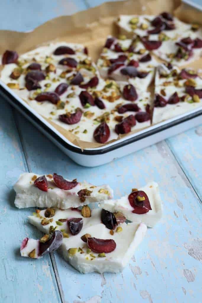 Frozen yogurt bark with cherries, pistachios and lemon zest in a white, parchment paper lined baking sheet with a black rim - a few pieces of bark are broken off and are set on the surface in front of the baking sheet