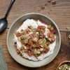 a bowl of honeyed yogurt topped with roasted rhubarb and hazelnut dukkah in the center - a sage green cloth, a dark colored spoon, and a small bowl of additional dukkah surrounding the bowl