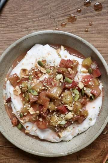 a bowl of honeyed yogurt topped with roasted rhubarb and hazelnut dukkah in the center - a sage green cloth, a dark colored spoon, and a small bowl of additional dukkah surrounding the bowl