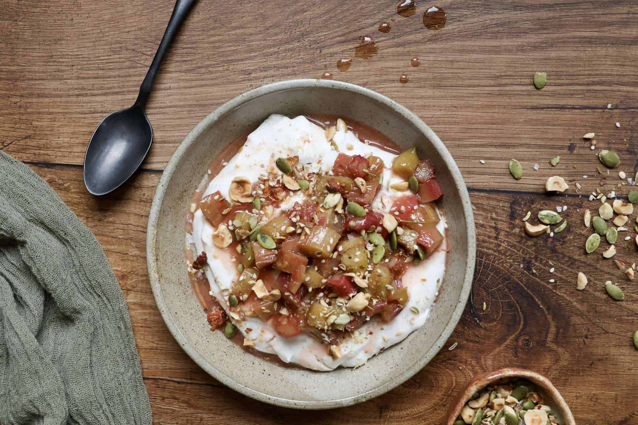 a bowl of honeyed yogurt topped with roasted rhubarb and hazelnut dukkah in the center - a sage green cloth, a dark colored spoon, and a small bowl of additional dukkah surrounding the bowl