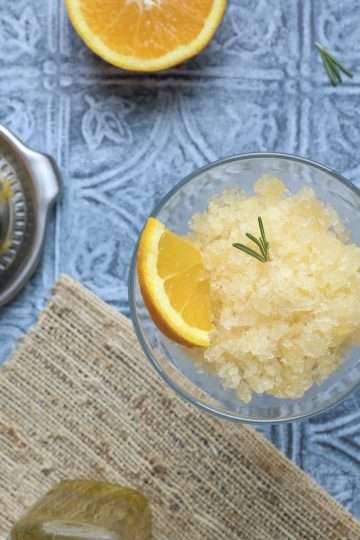 A cocktail glass filled with citrus granita, a small rosemary sprig, and a small orange triangle - a dark spoon is in the front and a couple small rosemary sprigs, half an orange, a small bottle of olive oil, and a silver orange juicer with half an orange are in the background