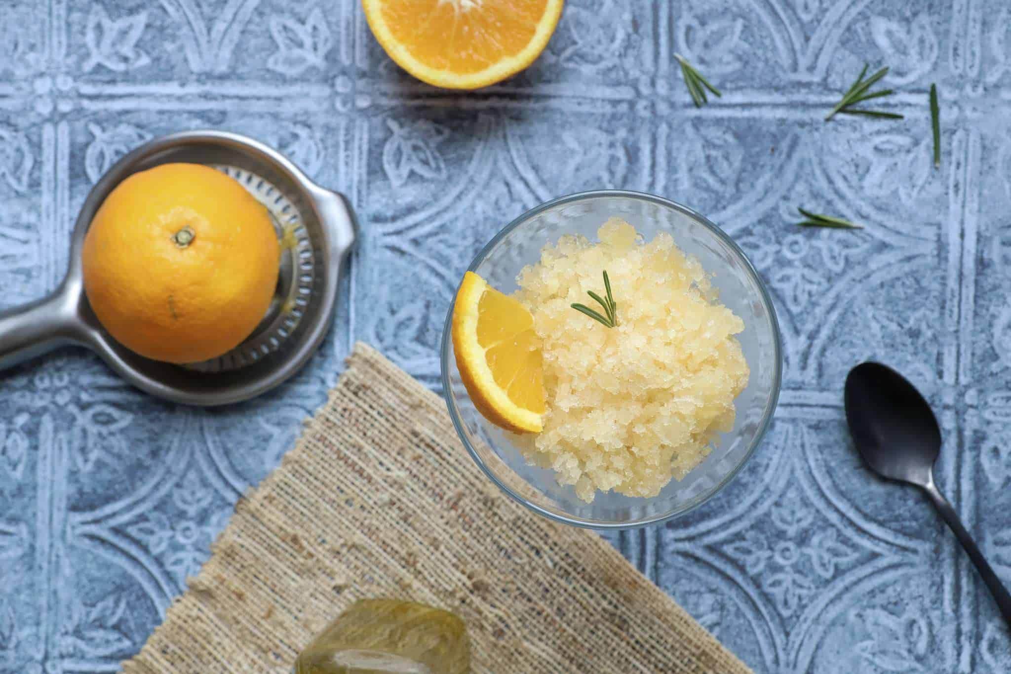 A cocktail glass filled with citrus granita, a small rosemary sprig, and a small orange triangle - a dark spoon is in the front and a couple small rosemary sprigs, half an orange, a small bottle of olive oil, and a silver orange juicer with half an orange are in the background