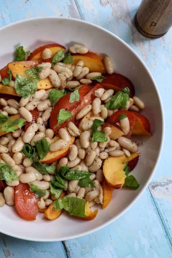 tomato, peach, basil and white bean salad in a white bowl and a pepper grinder can be partially seen in the background